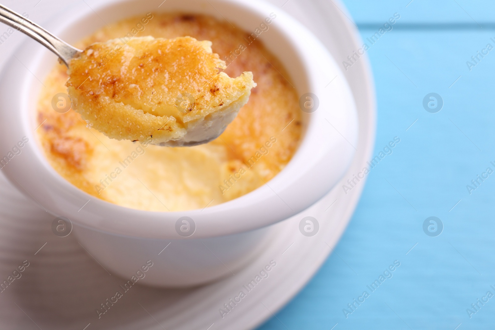 Photo of Taking delicious creme brulee with spoon from bowl at light blue table, closeup