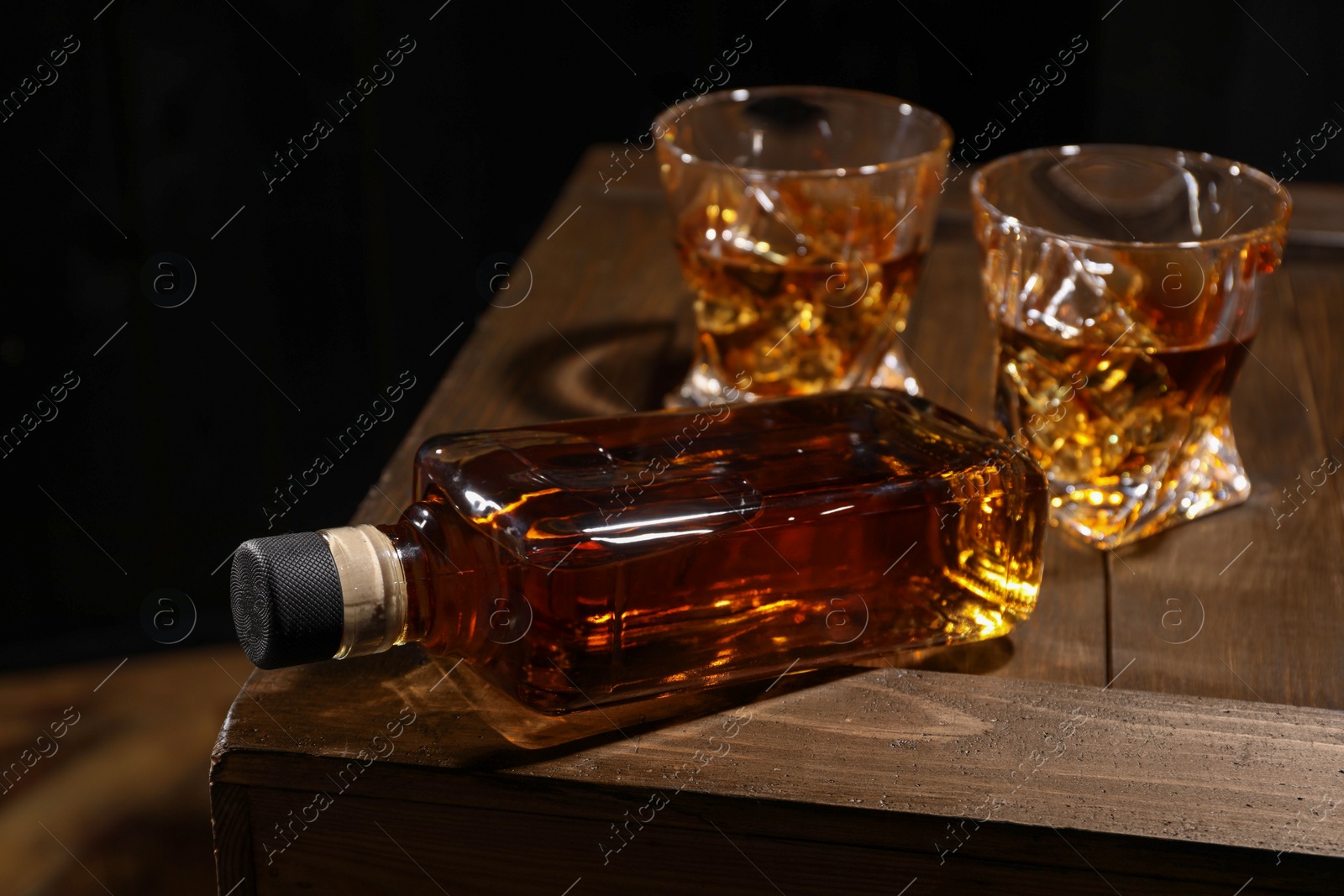 Photo of Whiskey with ice cubes in glasses and bottle on wooden crate against black background, closeup