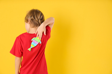 Photo of Little boy with paper fish on back against yellow background, space for text. April fool's day