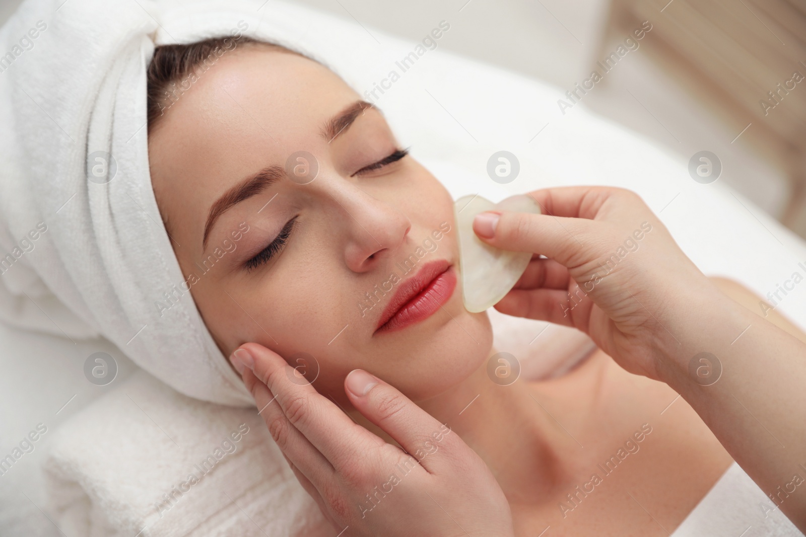 Photo of Young woman receiving facial massage with gua sha tool in beauty salon