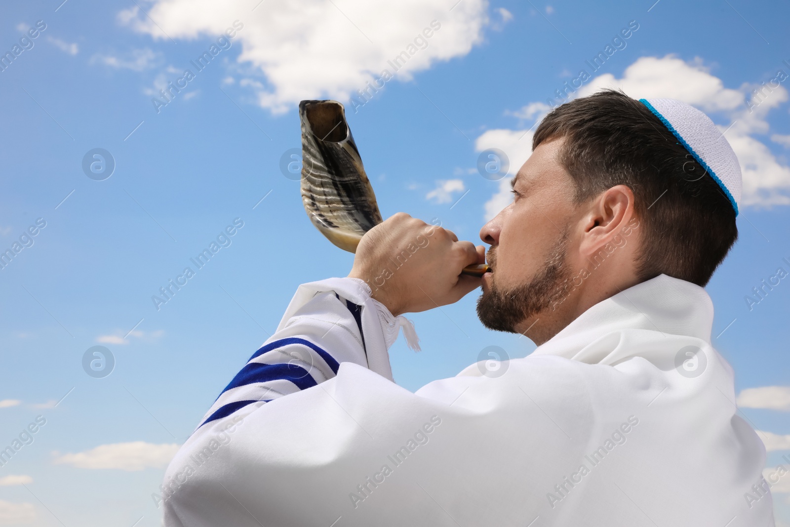 Photo of Jewish man in kippah and tallit blowing shofar outdoors. Rosh Hashanah celebration