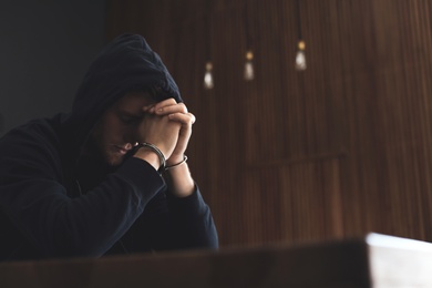 Man detained in handcuffs at table indoors, space for text. Criminal law