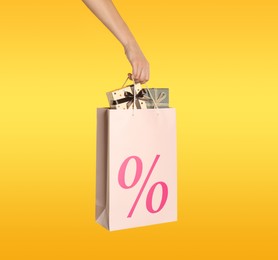 Woman holding paper shopping bag with percent sign on yellow background, closeup. Discount concept