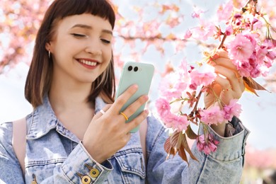Photo of Beautiful young woman taking picture of blossoming sakura tree branch in park, focus on hand