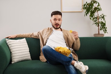 Handsome young man eating tasty potato chips on sofa at home