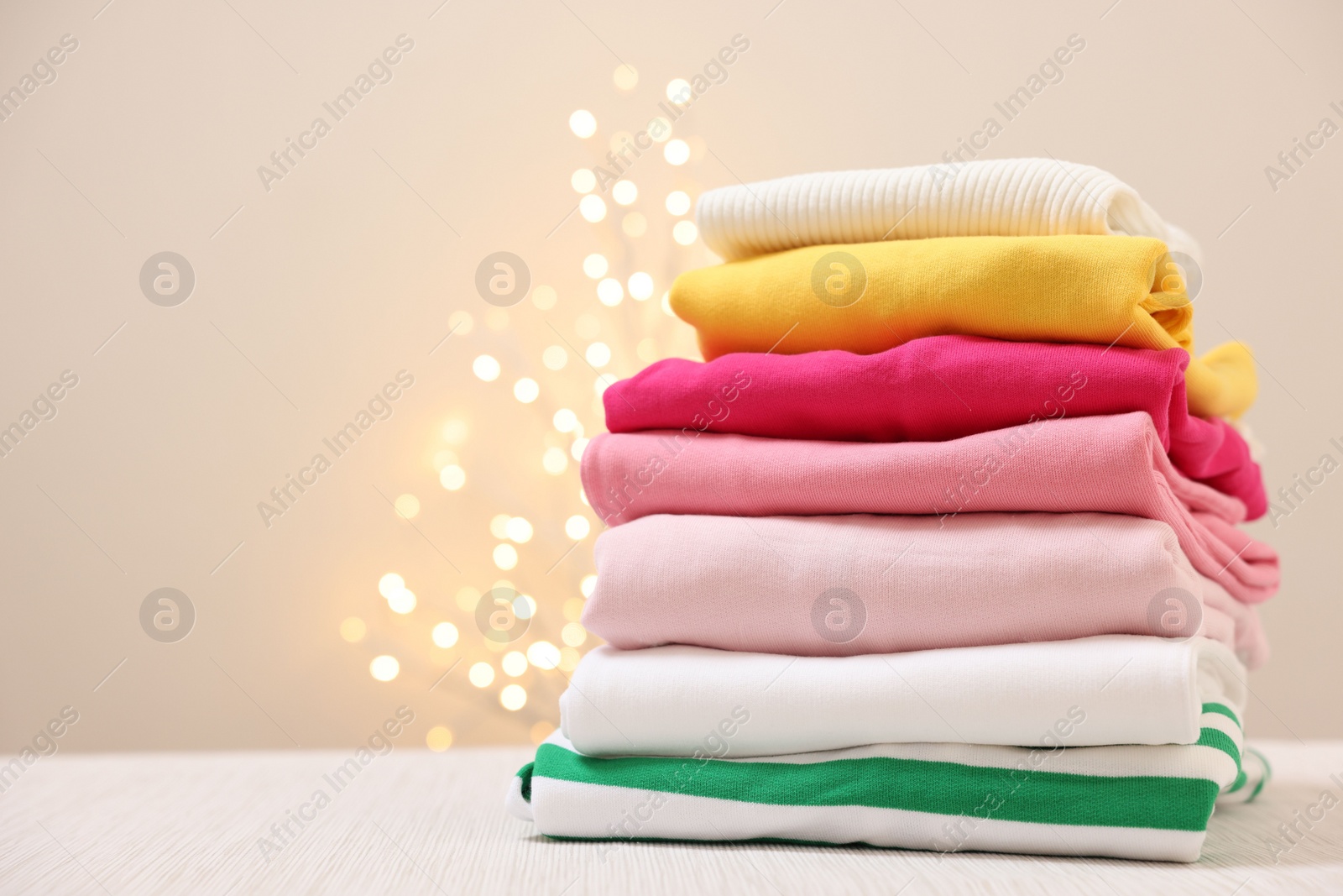 Stack of folded clothes on wooden table against beige background with blurred lights, space for text Photo of Stack of folded clothes on wooden table against beige background with blurred lights, space for text