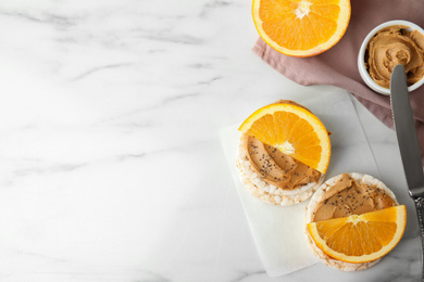 Photo of Puffed rice cakes with peanut butter and orange on white marble table, flat lay. Space for text