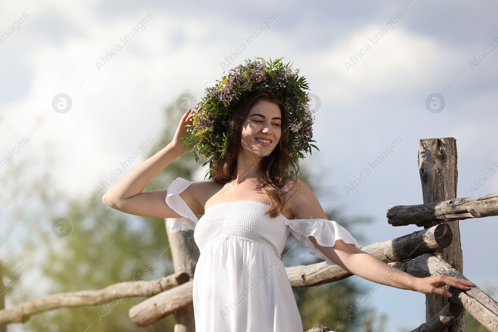 Photo of Young woman wearing wreath made of beautiful flowers near wooden fence on sunny day