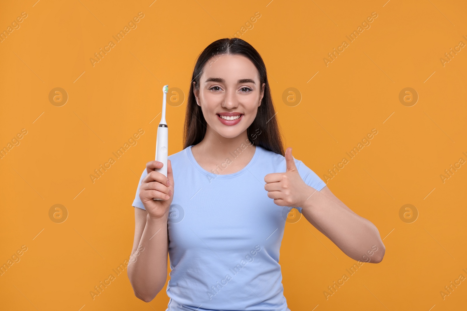 Photo of Happy young woman holding electric toothbrush and showing thumb up on yellow background