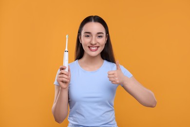 Happy young woman holding electric toothbrush and showing thumb up on yellow background