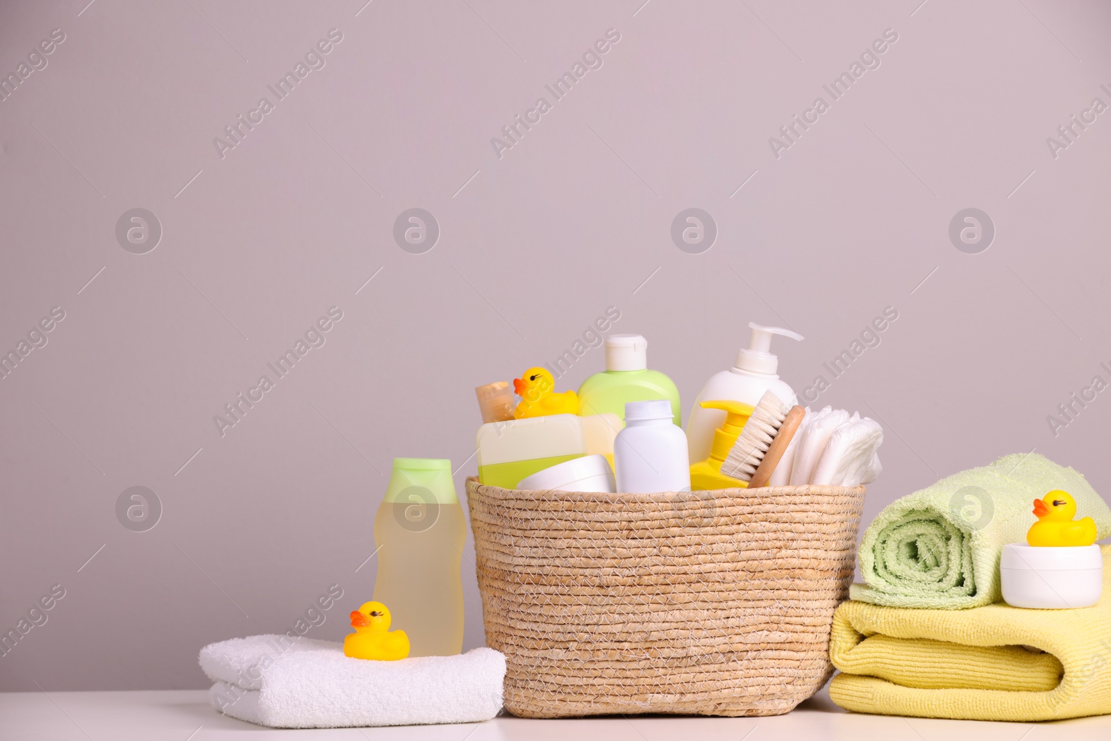 Photo of Wicker basket with baby cosmetic products, bath accessories and rubber ducks on white table against grey background. Space for text