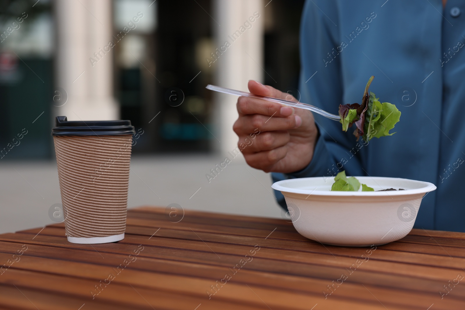 Photo of Businesswoman with plastic bowl of salad and paper cup of coffee having lunch at wooden table outdoors, closeup