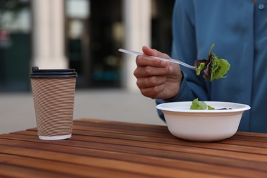 Photo of Businesswoman with plastic bowl of salad and paper cup of coffee having lunch at wooden table outdoors, closeup