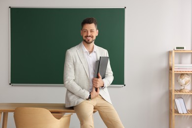 Photo of Happy teacher with stationery at desk in classroom