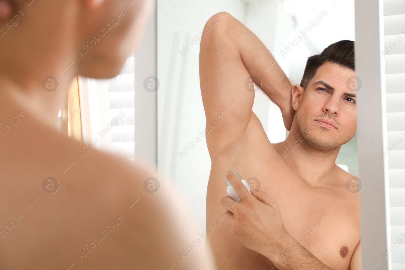 Photo of Handsome man applying deodorant to armpit near mirror at home