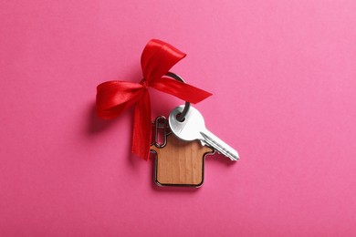 Key with trinket in shape of house and bow on pink background, top view. Housewarming party
