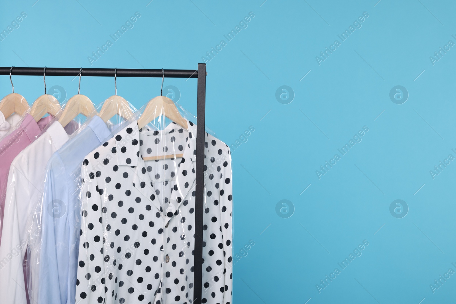 Photo of Dry-cleaning service. Many different clothes in plastic bags hanging on rack against light blue background, space for text