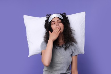 Photo of Tired young woman with sleep mask and pillow yawning on purple background. Insomnia problem