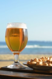 Photo of Glass of cold beer and pistachios on wooden table near sea
