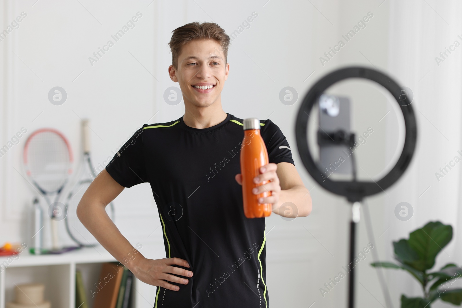 Photo of Smiling sports blogger holding bottle while streaming online fitness lesson with smartphone at home