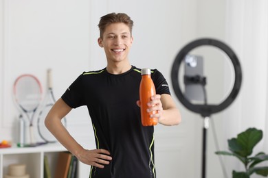 Smiling sports blogger holding bottle while streaming online fitness lesson with smartphone at home