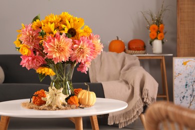 Photo of Beautiful bouquet with bright flowers and small pumpkins on white coffee table near sofa in room, space for text. Autumn atmosphere