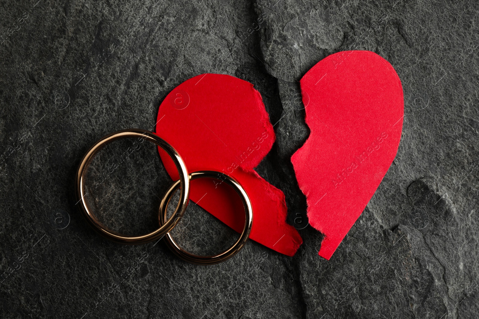 Photo of Halves of torn red paper heart and wedding rings on dark grey table, top view. Broken heart