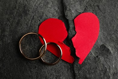 Photo of Halves of torn red paper heart and wedding rings on dark grey table, top view. Broken heart