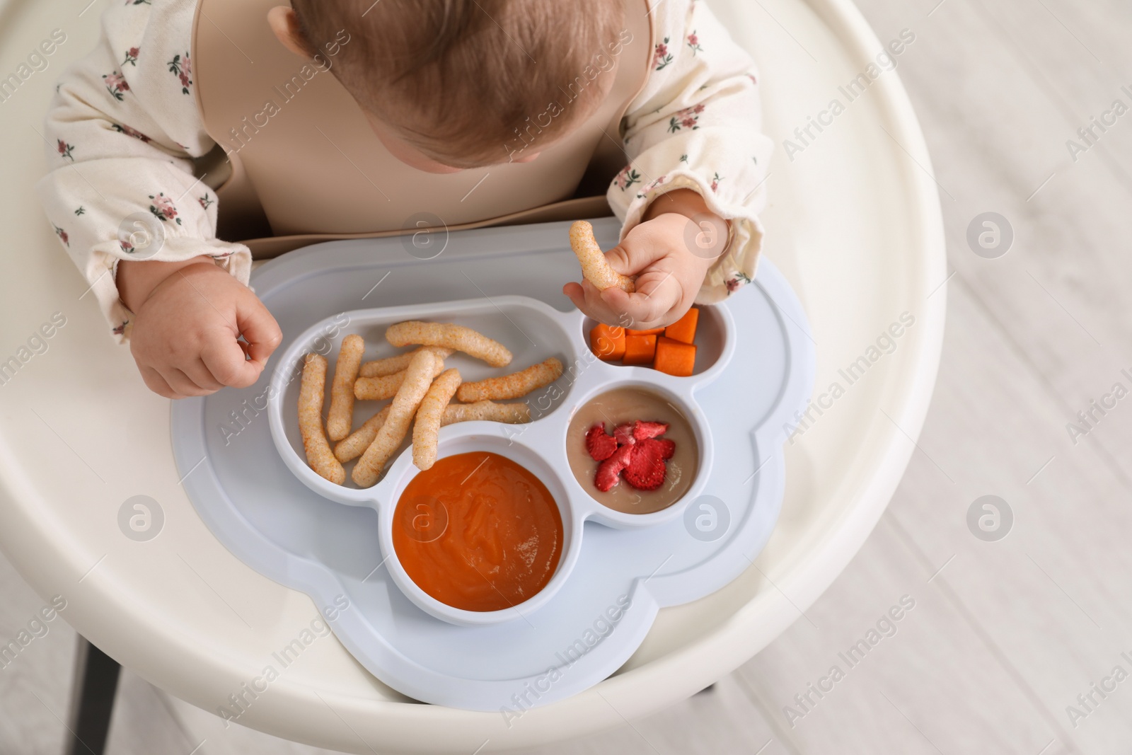 Photo of Little baby eating food in high chair indoors, closeup. Above view