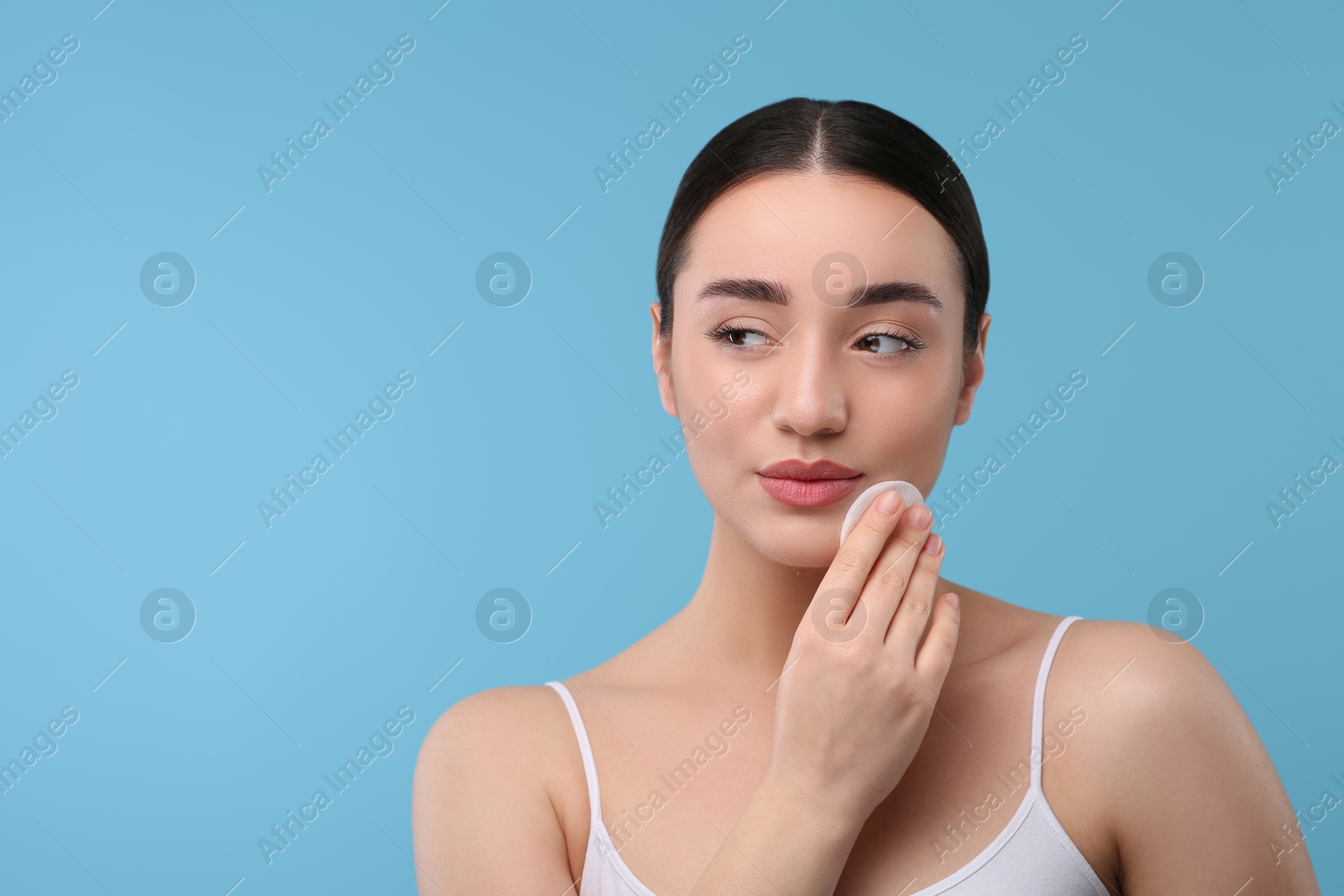 Photo of Beautiful woman removing makeup with cotton pad on light blue background, space for text