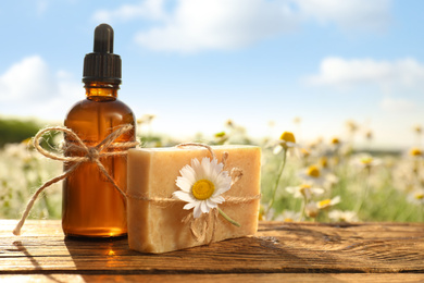 Photo of Bottle of chamomile essential oil and soap bar on wooden table in field