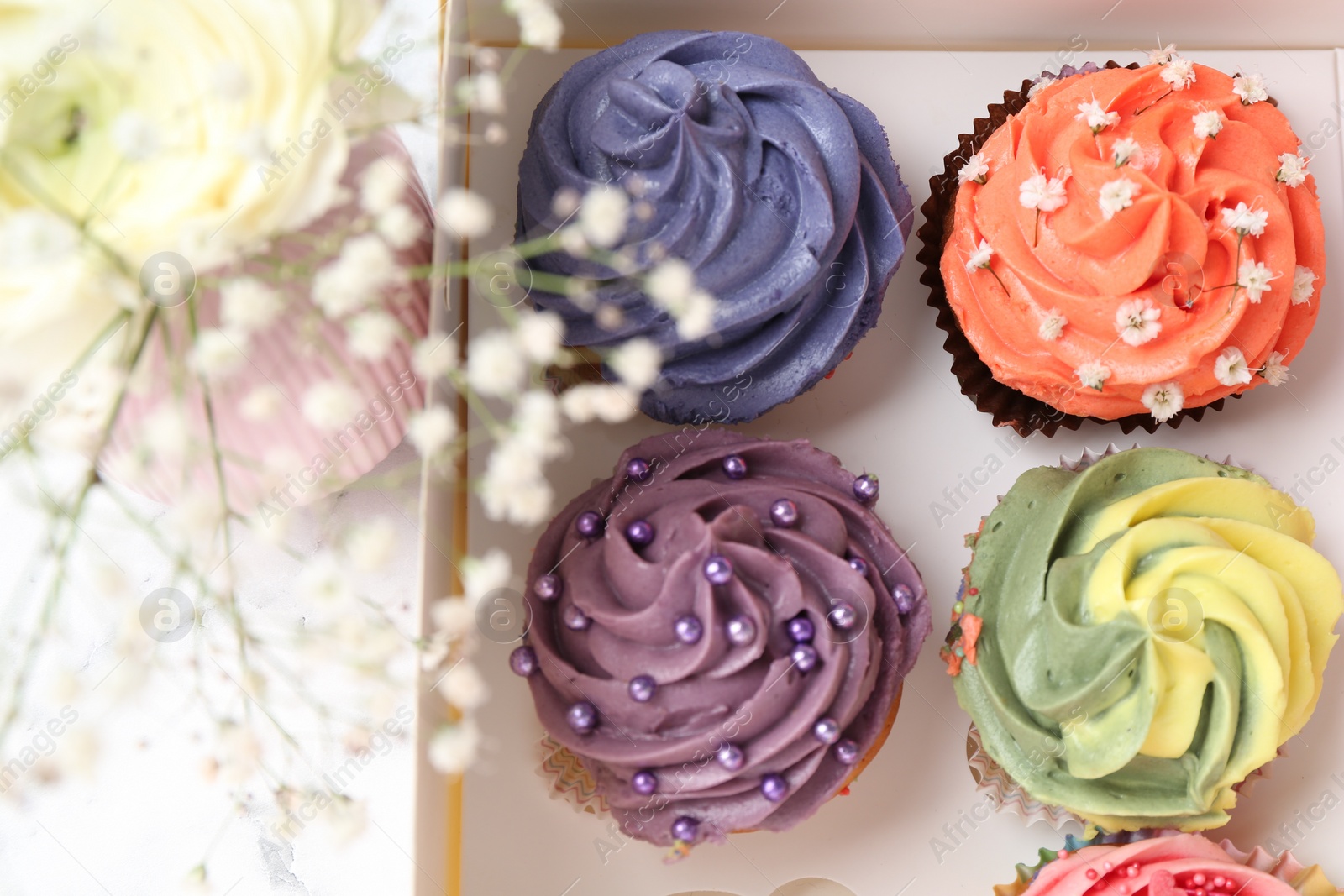 Photo of Different colorful cupcakes in box and flowers on table, closeup