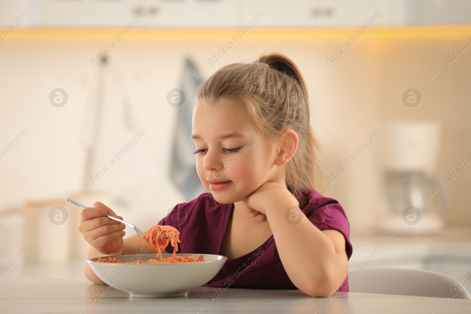 Photo of Cute little girl eating tasty pasta at table in kitchen