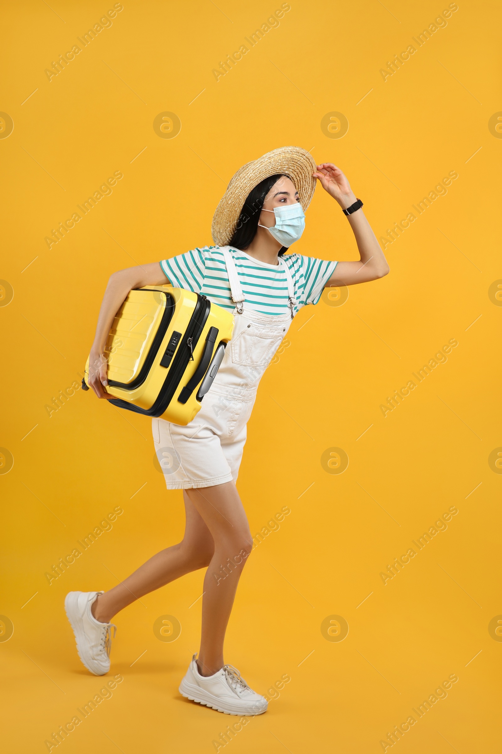 Photo of Female tourist in medical mask with suitcase on yellow background. Travelling during coronavirus pandemic