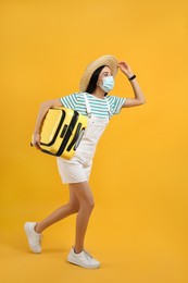 Female tourist in medical mask with suitcase on yellow background. Travelling during coronavirus pandemic