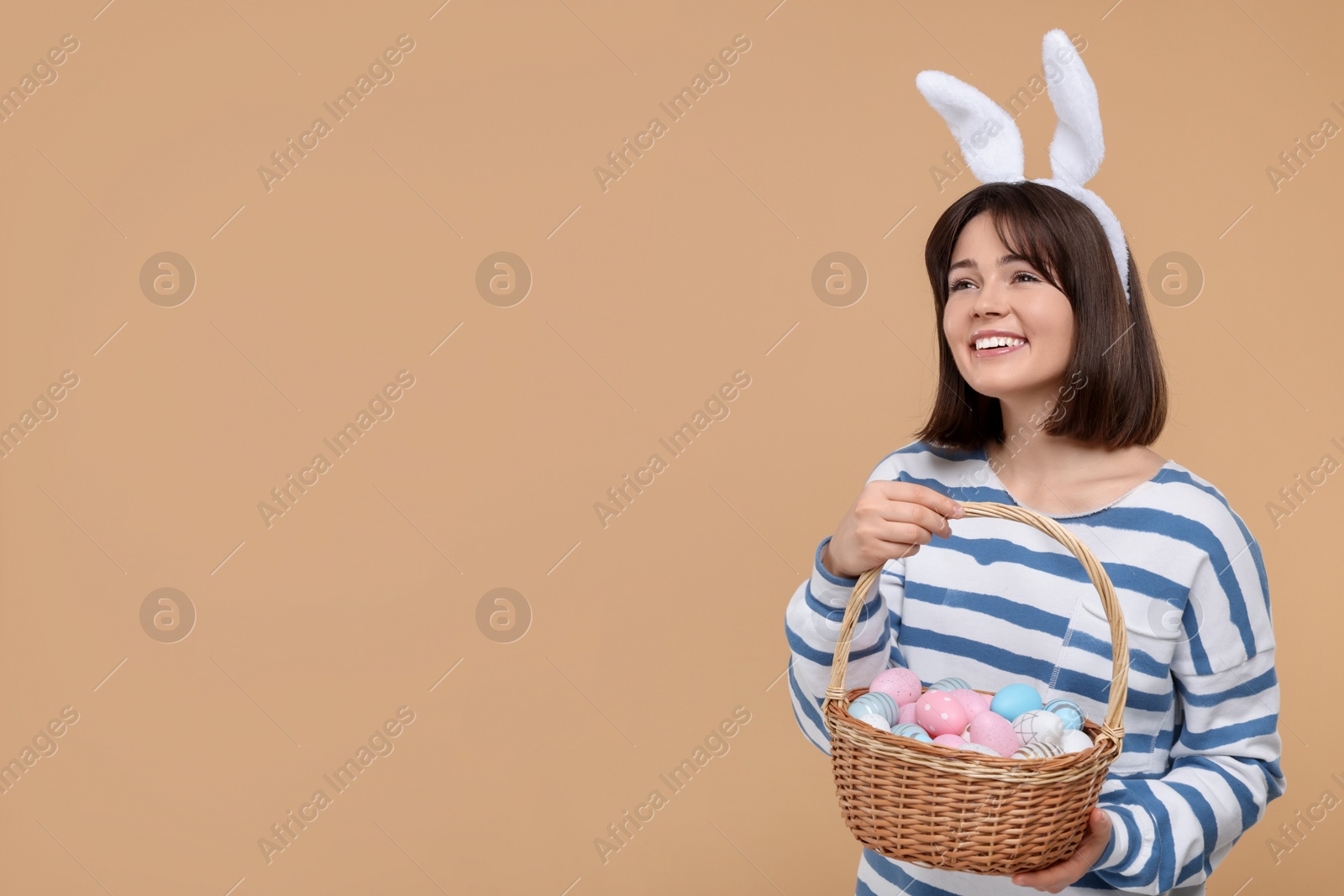 Photo of Easter celebration. Happy woman with bunny ears and wicker basket full of painted eggs on beige background, space for text