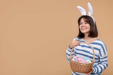 Photo of Easter celebration. Happy woman with bunny ears and wicker basket full of painted eggs on beige background, space for text