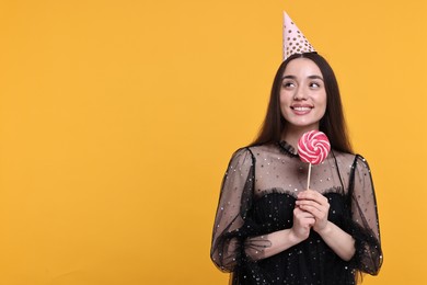 Photo of Happy woman in party hat holding lollipop on orange background, space for text