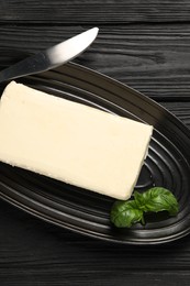 Photo of Block of tasty butter with basil and knife on black wooden table, top view