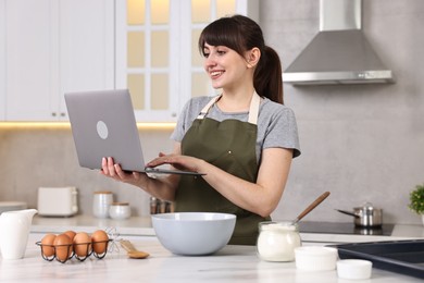 Photo of Happy young housewife using laptop while cooking at white marble table in kitchen