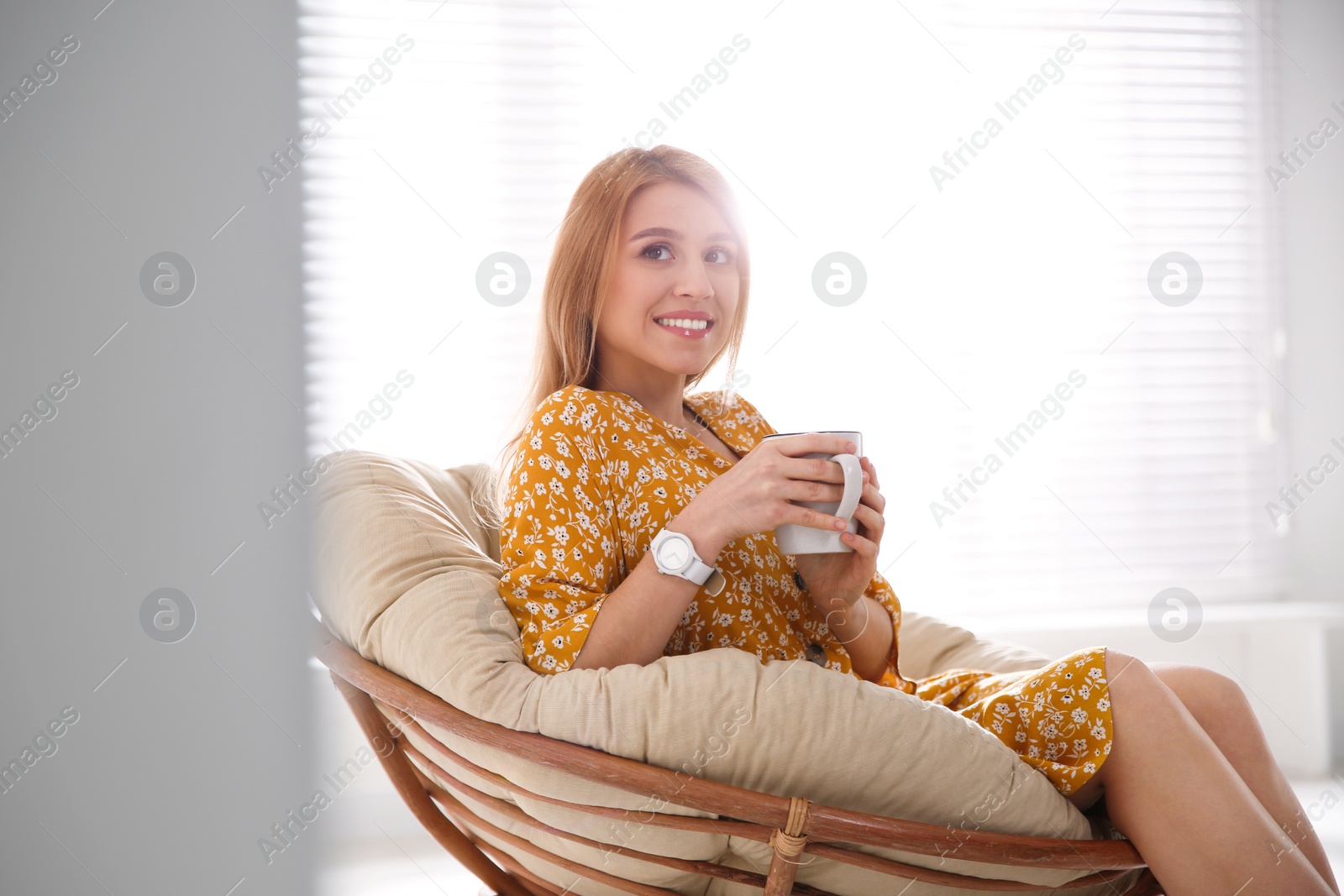 Photo of Young woman with cup of drink relaxing in papasan chair near window at home