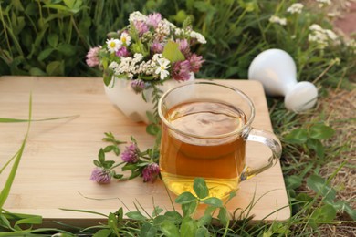 Cup of aromatic herbal tea, pestle and ceramic mortar with different wildflowers on wooden board in meadow