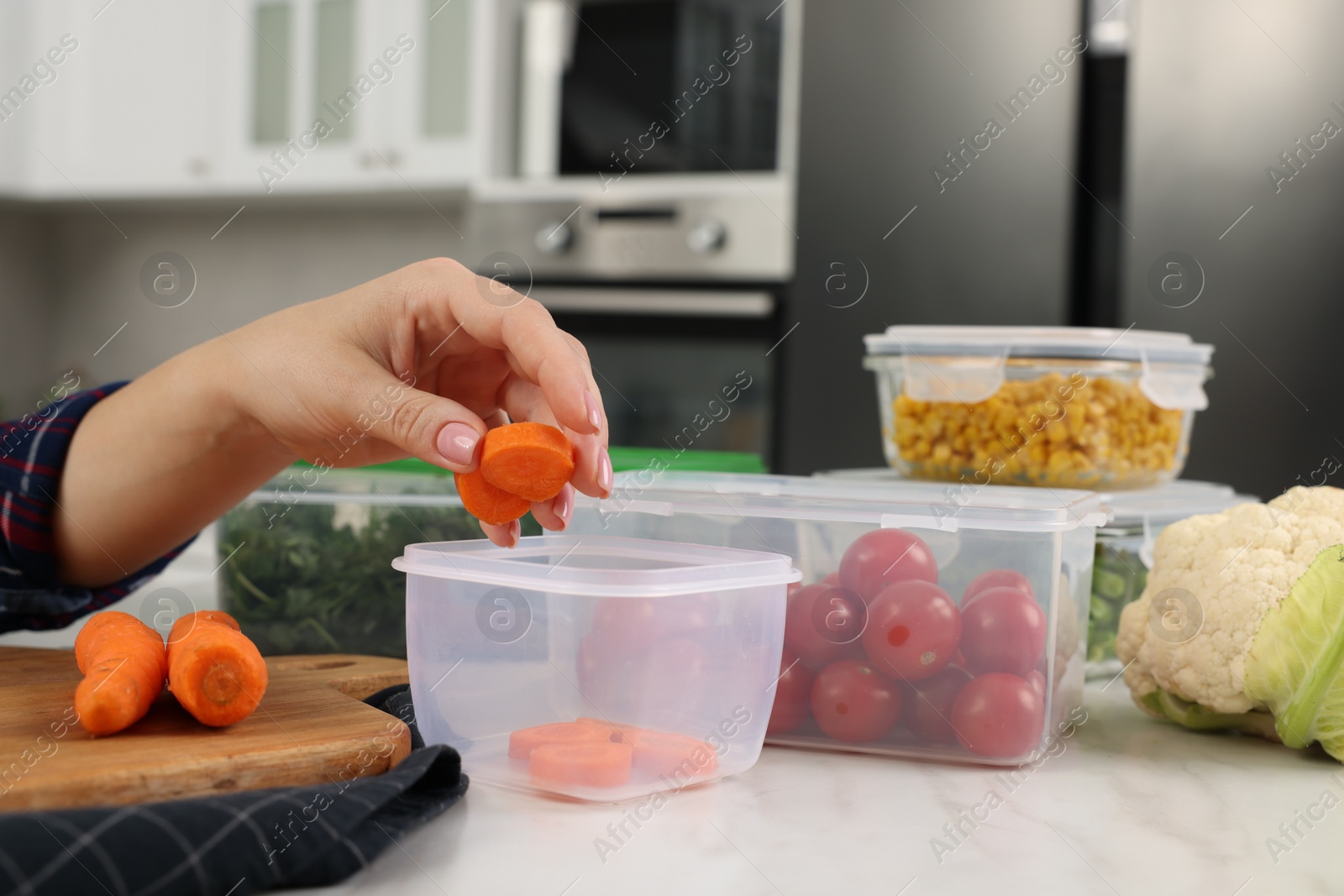 Photo of Woman putting fresh carrot into plastic container at white marble table, closeup. Food storage