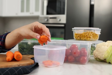 Woman putting fresh carrot into plastic container at white marble table, closeup. Food storage