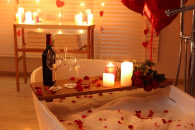 Photo of Wooden tray with wine, burning candles and rose petals on tub in bathroom. Valentine's day celebration