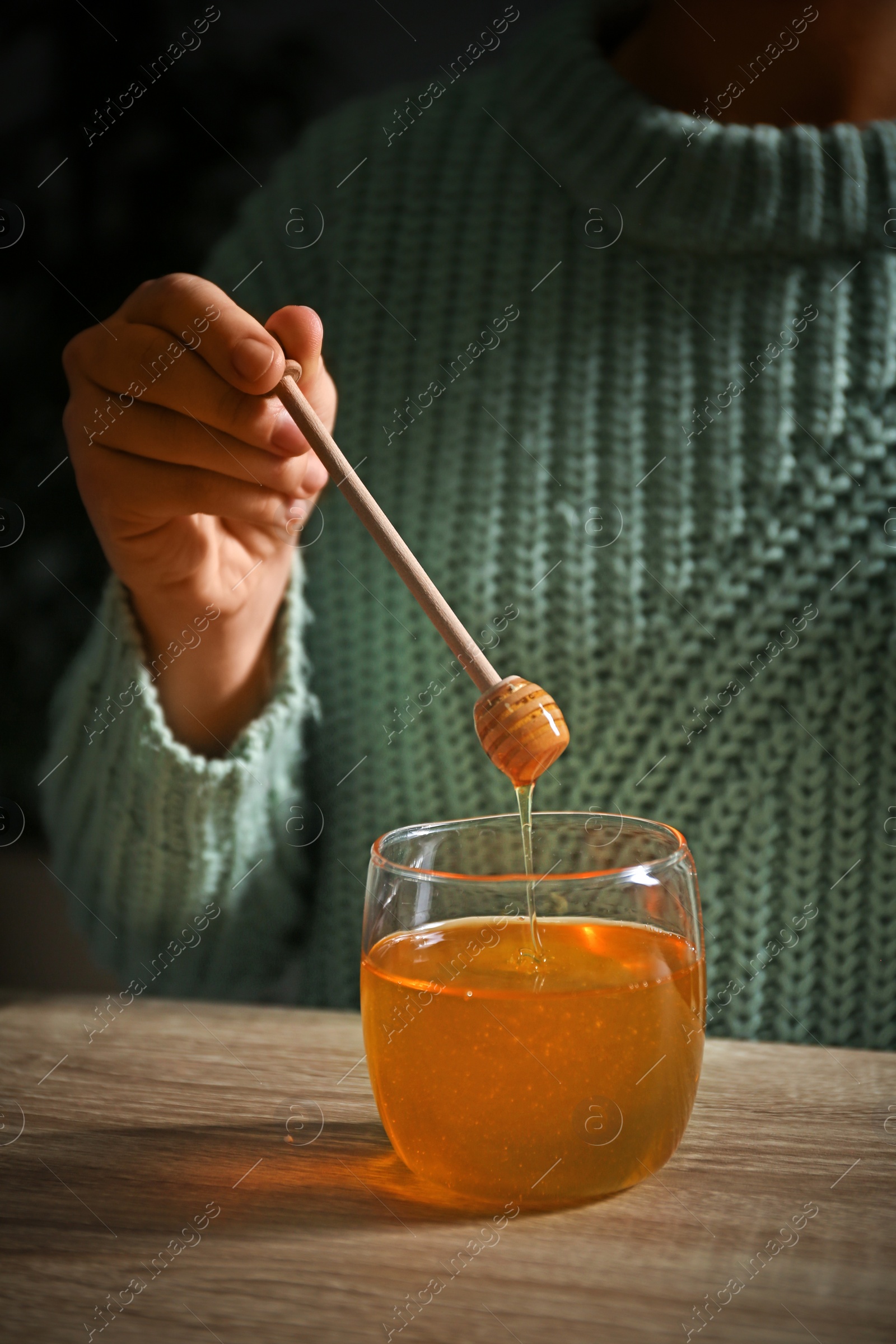 Photo of Woman holding honey dipper over jar at wooden table, closeup