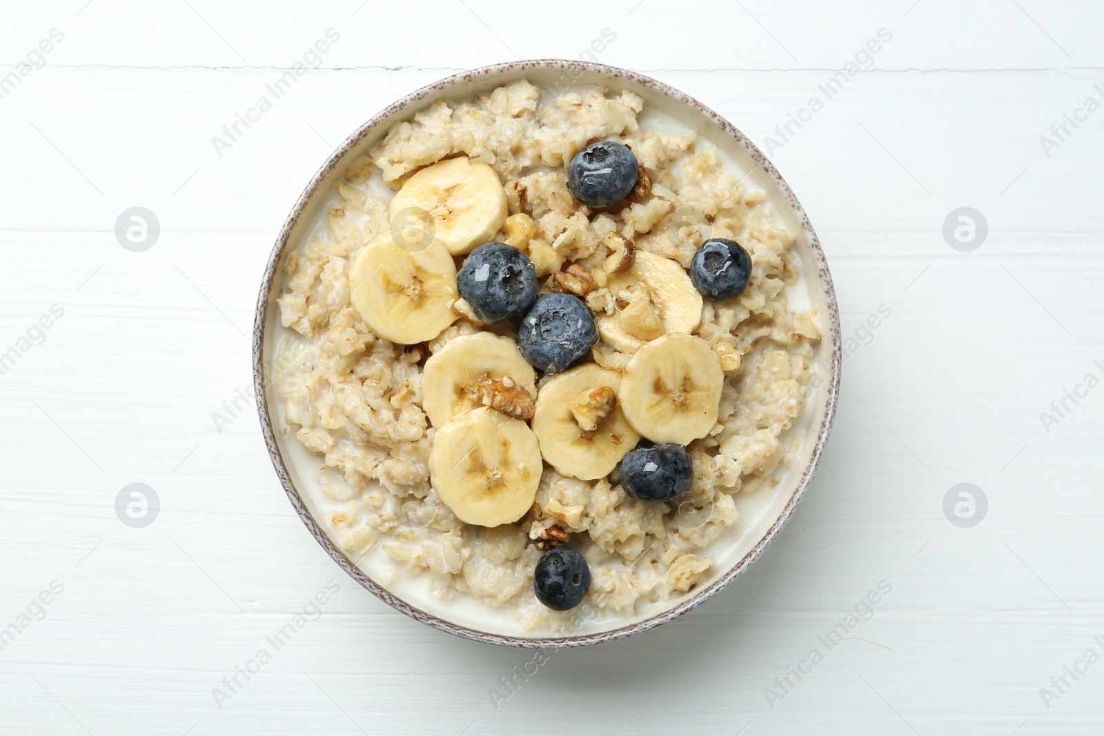 Photo of Tasty oatmeal with banana, blueberries, walnuts and honey served in bowl on white wooden table, top view