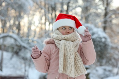 Photo of Cute little girl wearing Santa hat in snowy park on winter day