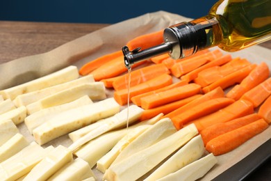 Photo of Pouring oil onto baking tray with parsnips and carrots against blue background, closeup
