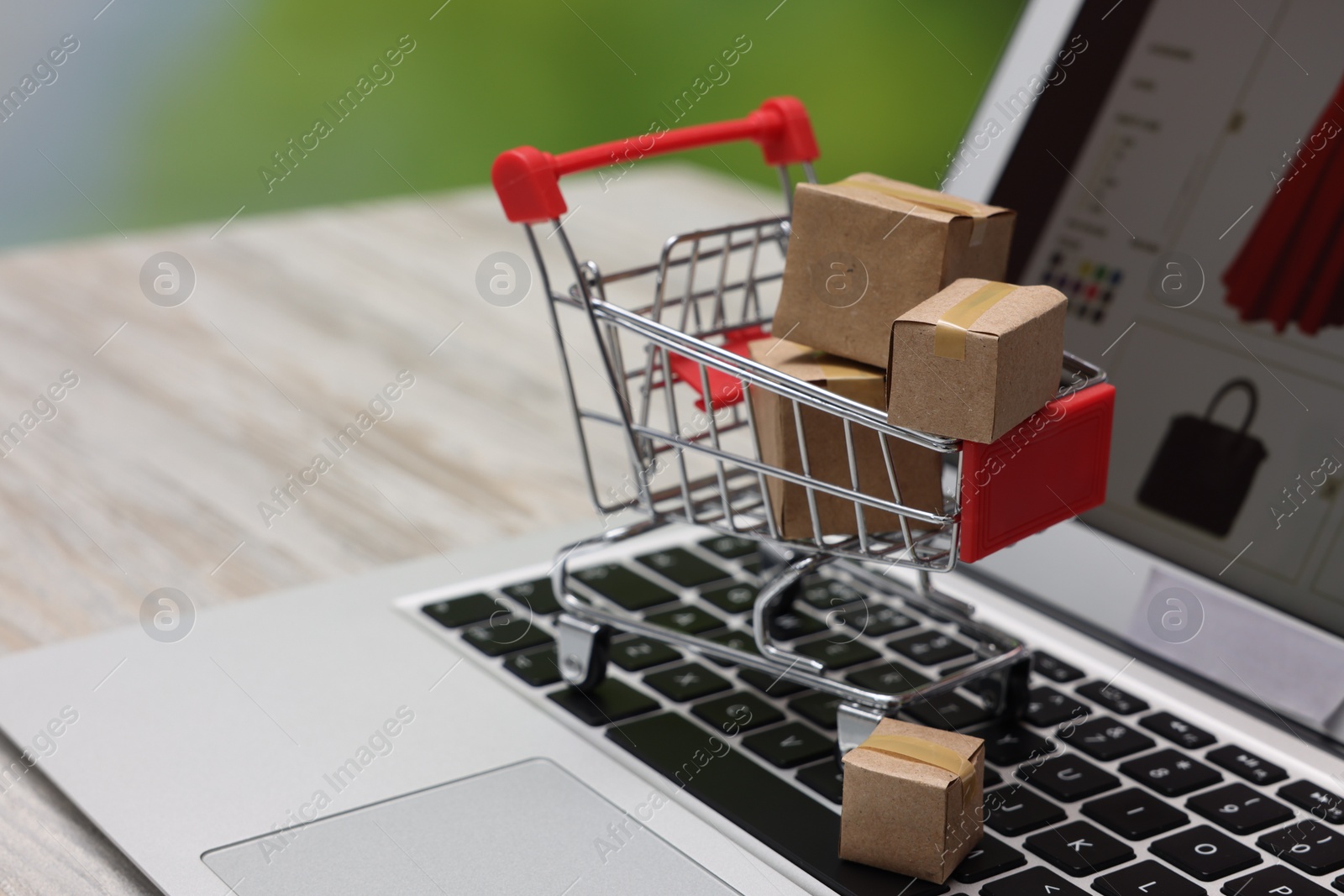 Photo of Internet store. Small cardboard boxes, shopping cart and laptop on light wooden table, closeup. Space for text